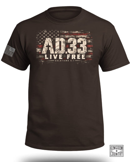 A dark brown crewneck t-shirt featuring a distressed American flag graphic, the text AD 33, Live Free, and Galatians 5:1.