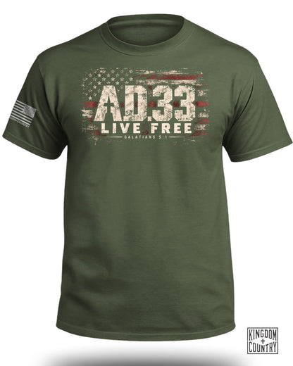 An olive green t-shirt featuring a distressed American flag graphic, the text AD.33 LIVE FREE, and Galatians 5:1.