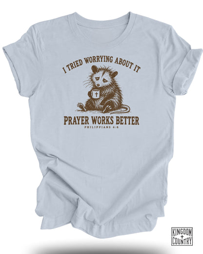Light blue t-shirt with an opossum holding a mug, text "I tried worrying about it prayer works better".